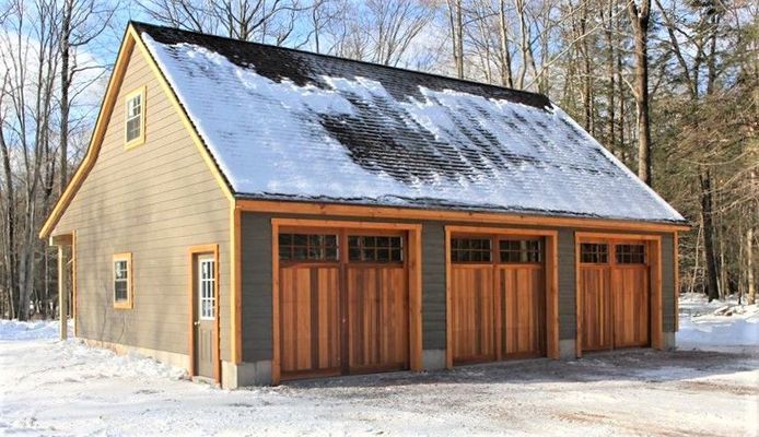 Three-bay garage with wood doors and snow-covered roof in a snowy, wooded setting.