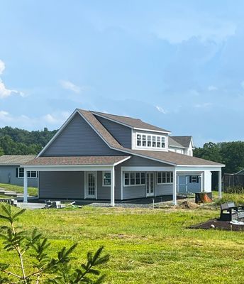 Blue-gray house with white trim and a large porch, set in a grassy field with a partly cloudy sky.