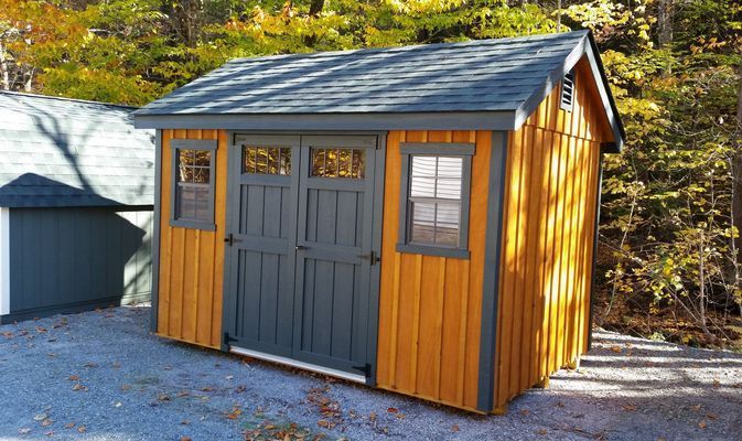 Wooden shed with gray door and trim, windows, blue roof, set outdoors near trees.