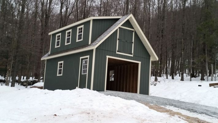 Green two-story barn in snow, with a gravel driveway leading into the open garage door.