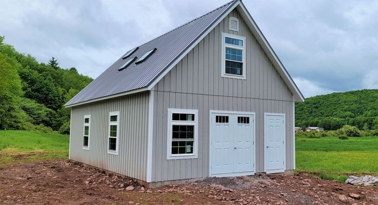 Gray two-story garage with a metal roof, windows, and a door, set in a grassy field against a hilly backdrop.