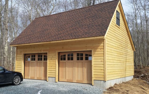 Two-car garage with wood siding, brown roof, and tan garage doors. Car parked beside it, trees in background.