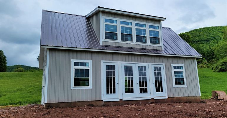 A light gray building with a metal roof, dormer windows, and French doors in a grassy field.