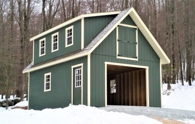 Green two-story barn-style building with white trim, open garage, and small windows, set in a snowy wooded area.