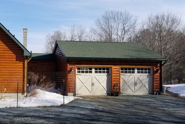 Two-car garage with white doors and a green roof, next to a log cabin on a sunny day with snow.
