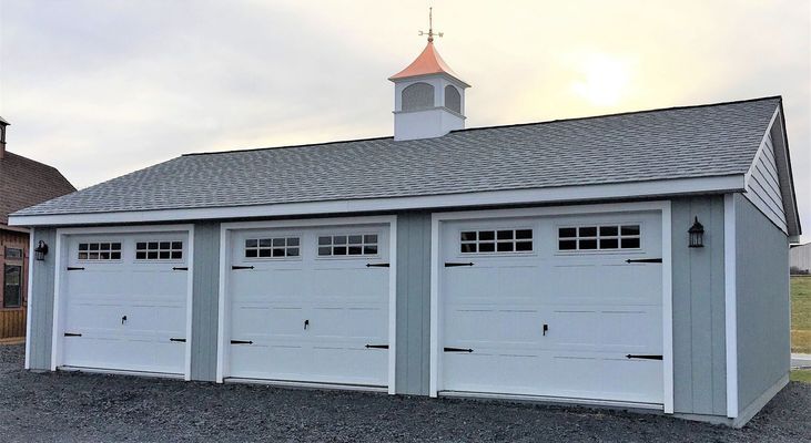 Three-car garage with white doors, light blue siding, and a cupola, set against a cloudy sky.