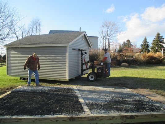 Two men moving a shed with a machine onto a gravel foundation in a yard.