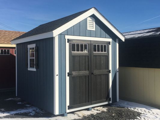 Blue shed with dark gray doors and white trim, with a window and black roof. Snow is on the ground.