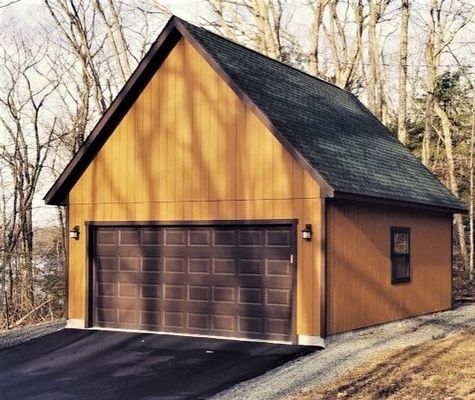 Brown garage with dark green roof, brown door, and small window.  Set on a paved driveway with a wooded background.