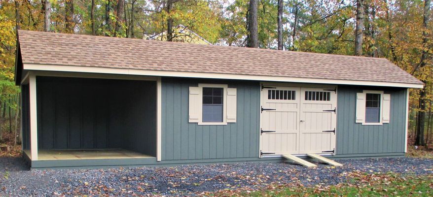 A long, blue shed with a covered open side and a garage door; set in a wooded area.