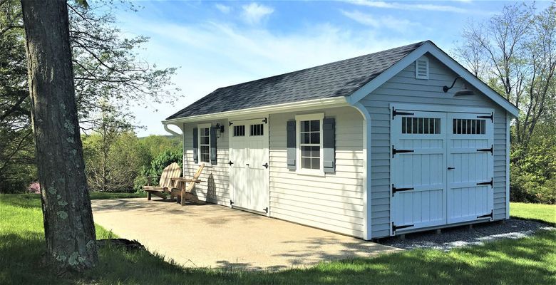 Blue-sided shed with white garage doors and shutters, on concrete slab, trees and grass in the background.