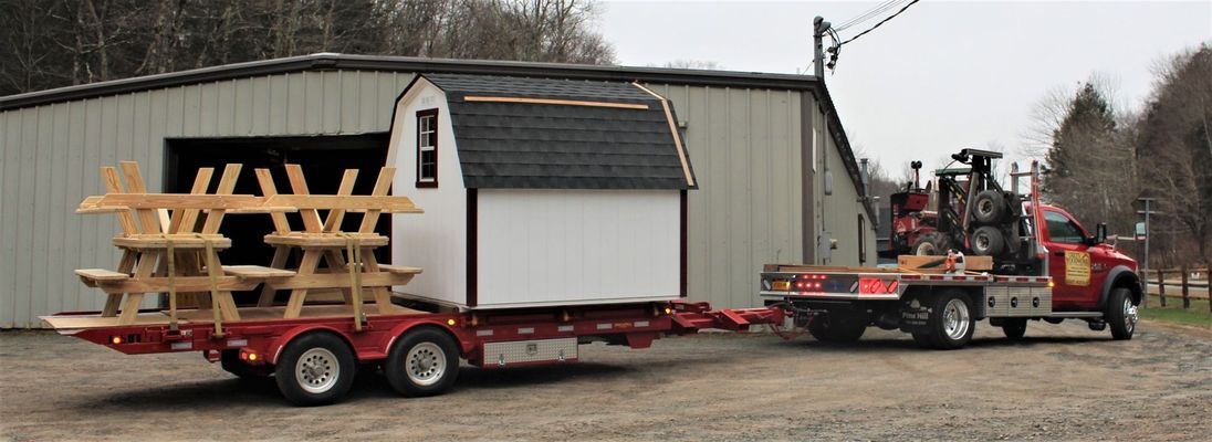 A red flatbed truck towing a trailer carrying a shed and picnic tables in front of a large building.