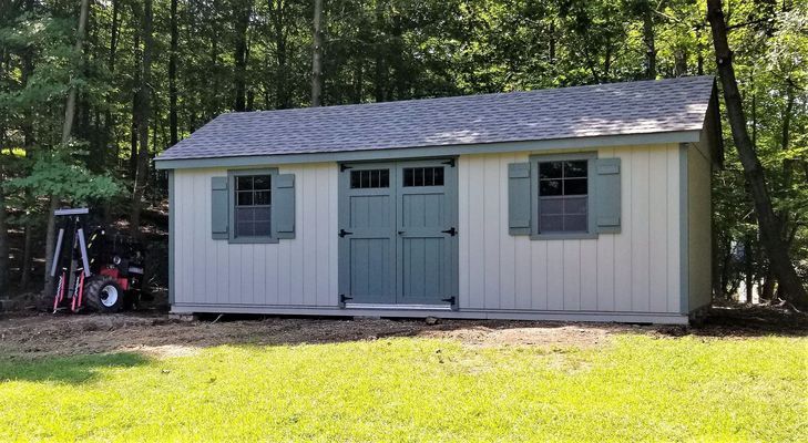 Tan shed with teal door, shutters, and trim. Set in a yard with a lawn and forest backdrop.