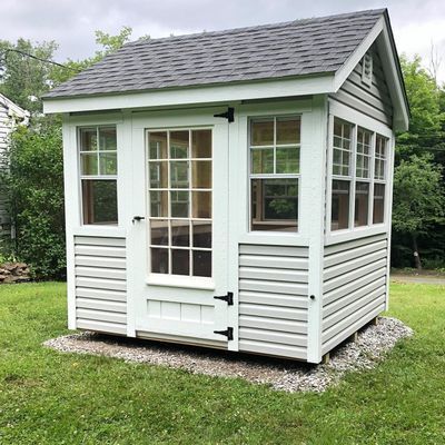 Small, white shed with windows and door on gravel foundation, set in grassy yard.