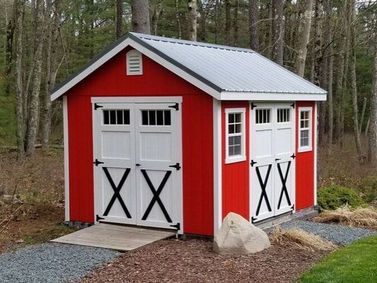 Red shed with white doors, black accents, and a gray roof, set in a wooded area.
