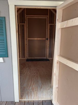 Open doorway reveals a small, unfinished room with plywood walls and floor. A screen door is visible.