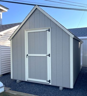 Gray shed with white trim and door, set on concrete blocks, against a blue sky.