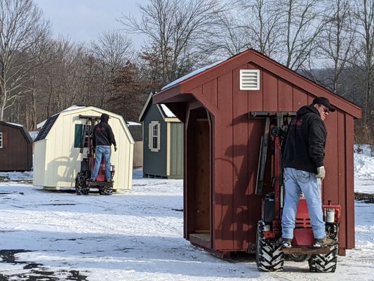 Two men on small tractors by sheds in a snowy field; red, beige and green sheds are visible.