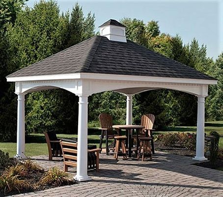 Gazebo with a dark roof, white columns, and wooden furniture on a brick patio in a garden.