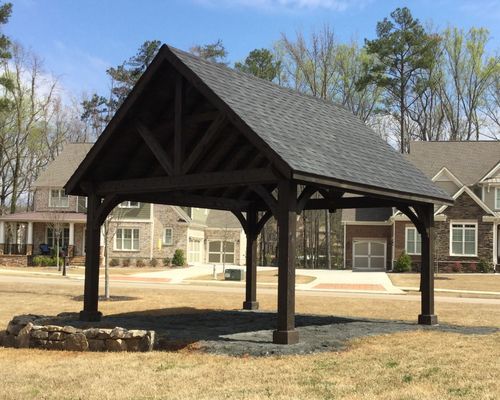 Dark brown gazebo in a grassy area with houses in the background on a sunny day.