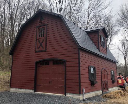 Red barn-style building with black roof, garage door, and dormer, set against a background of trees.