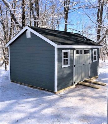 Blue shed with white trim, black roof, snow-covered ground, and trees in the background.