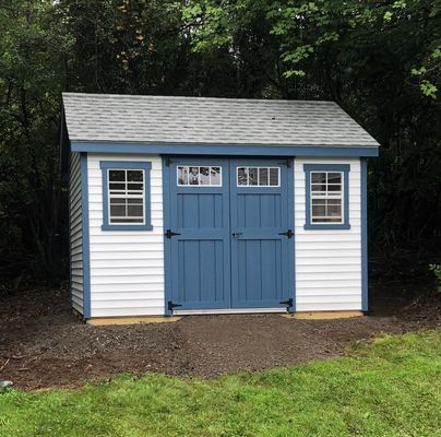 Wooden shed with black double doors and a blue shingle roof. Two windows on the sides.