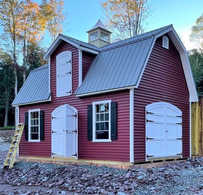 Red barn-style shed with white doors and shutters, gray roof, small cupola, and ladder beside it.