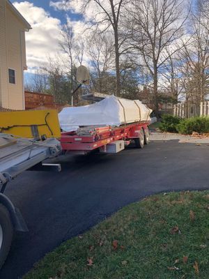 A trailer loaded with a large, white, boat-shaped object on a paved driveway, next to a house and trees.