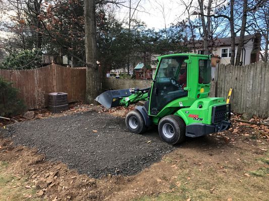 Green mini-loader on gravel, leveling a backyard area with a fence and trees.
