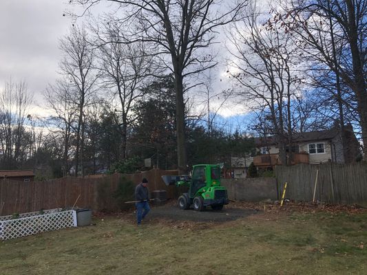 A person raking leaves near a green mini loader and a house with bare trees in the background.