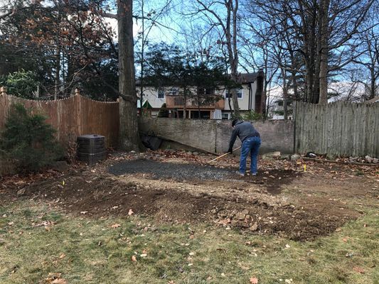 Person raking a small, dark patch of dirt in a backyard with brown fencing and a house in the background.