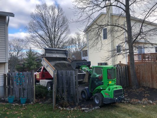 A green mini-loader dumps soil from a truck into a backyard. A house and fence are visible.