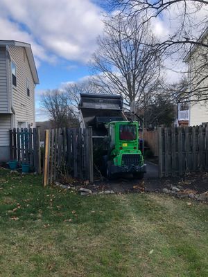 Green loader dumping contents into a backyard between a wooden fence and houses on a cloudy day.