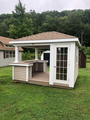 Small tan and white gazebo with a bar and a door, in a grassy yard with trees.