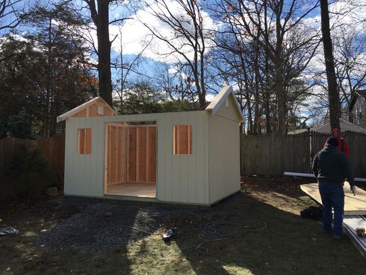 Shed under construction outdoors with a partially built roof, beige siding, and a person in the background.