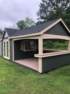 Green shed with bar area, beige trim, tan deck. French doors and window with folding shelf. Overcast day, green grass.