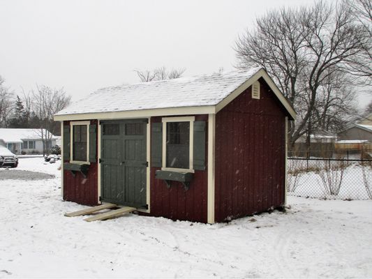 Red shed with snow on the roof and ground, green doors and shutters, in a wintery yard.
