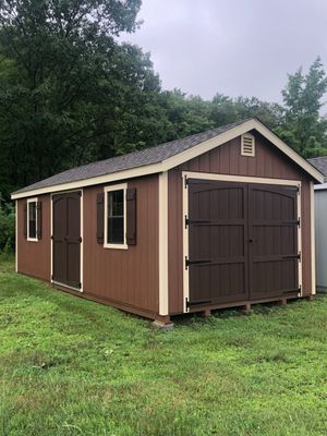Brown shed with cream trim, two windows, and a double door, set in a grassy yard with trees in the background.