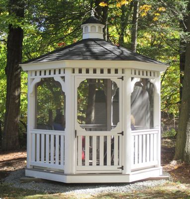 White octagonal gazebo with black roof in a wooded area.