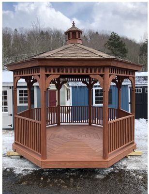 Brown octagonal gazebo with shingled roof, open-air with railings, set on a wooden deck, outdoors.