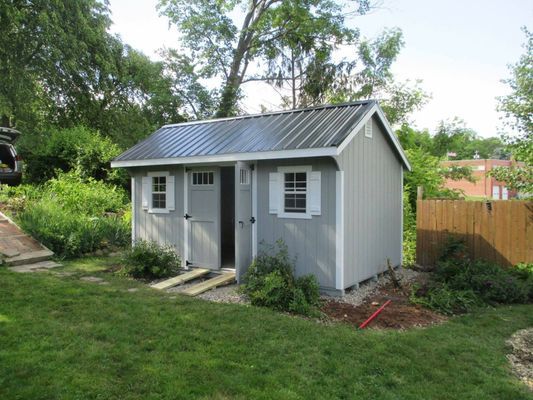Light grey shed with metal roof, open door, small window, surrounded by greenery, lawn in foreground.