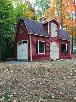 Red barn-style building with tan doors and trim, black shutters, and a cupola, set in a wooded area with fall foliage.