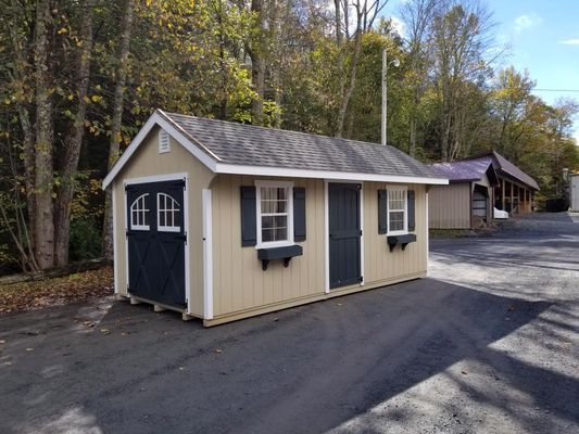 Tan shed with dark blue doors and shutters, set on a paved area, with trees in the background.