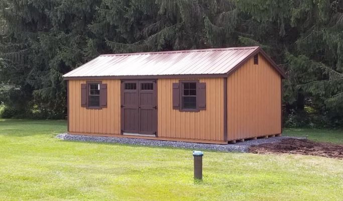 Brown shed with brown roof, doors, and shutters, sitting on gravel in a grassy yard.