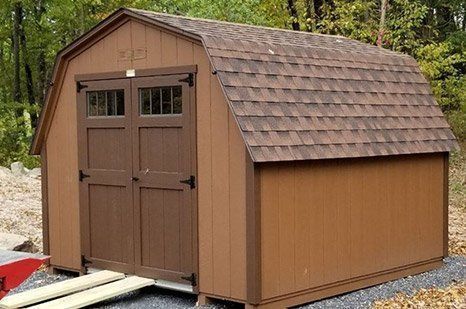 Brown wooden barn-style shed with double doors, dark roof, and brown trim; outdoors.