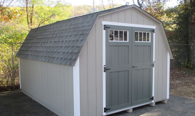 Tan shed with barn-style roof, grey doors, and white trim against a backdrop of trees.