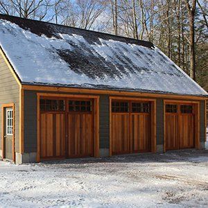 Three-bay wooden garage with snow-covered roof and doors.  Located in a wintery setting with trees.
