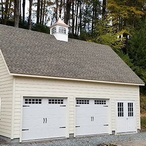 Beige two-car garage with white doors, a small cupola, and a light gray roof against a wooded background.
