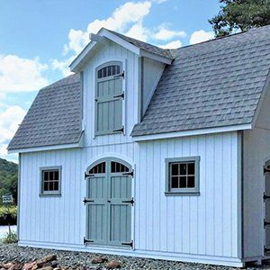 White shed with gray roof, doors, and trim, featuring a dormer and arched doorway, set against a blue sky.